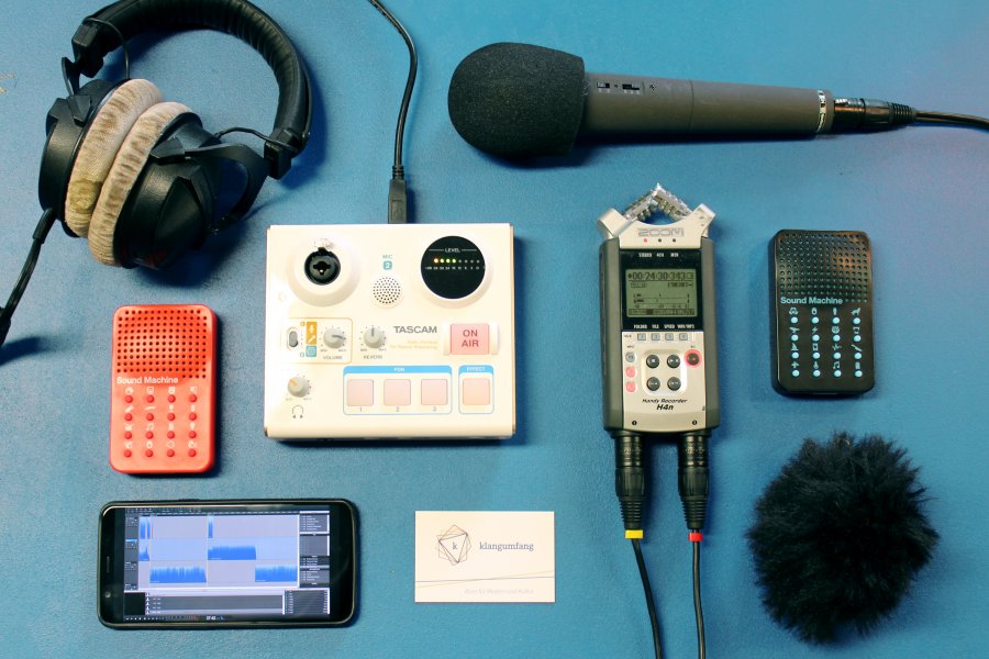 Auf einem blauen Tisch liegen Mikrofon, Aufnahmegerät, Kopfhörer und andere Audio-Spielereien.